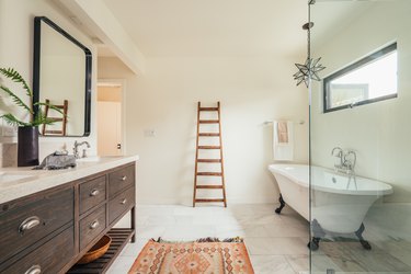 White bathroom with clawfoot tub, boho rug, wood dresser double sink and wood ladder against wall.