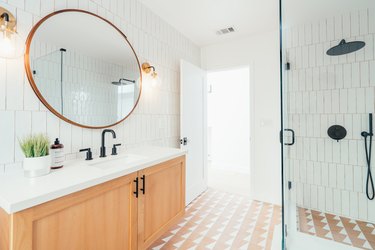 Bathroom with neutral-white floor tile, white vertical wall tile, round mirror, glass door shower, and light wood vanity