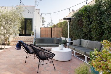 a backyard of a Spanish-style home with hexagon Terracotta pavers, two gray outdoor couches, two black patio chairs, a white, round coffee table, string lights, large privacy hedges