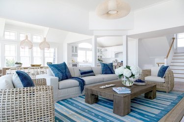 coastal living room with chairs, off-white sofa, blue striped area rug, blue throw pillows, white walls, wood floor.