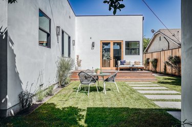 a gray house with a flat roof and a backyard with evenly spaced, modern concrete rectangular steps, various green plants and a table with three chairs