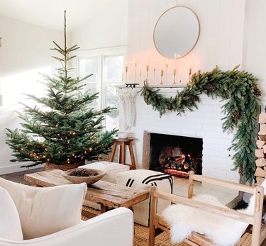 Christmas Tree Themes with Douglas fir Christmas tree, green garland on mantle, white brick fireplace, round mirror, candlesticks, white couch, sheepskin on side chair, rustic coffee table.