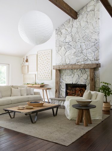 Stone farmhouse fireplace with wood beams, large Noguchi style pendant, rustic coffee table, end table, chair and couch.