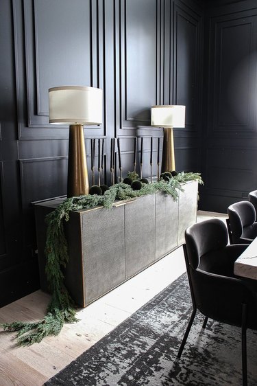 Modern Christmas decor with black walls, matching brass lamps, green garland, black and gray area rug, black dining chairs