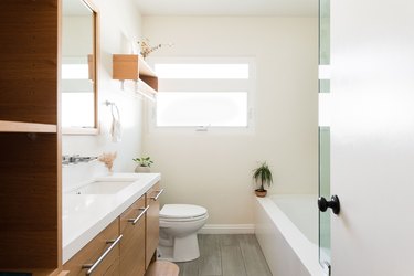 bathroom with wood vanity, wood flooring, white toilet, over-the-toilet wood shelf, bathtub with glass shower door, small plant on the ledge of the tub