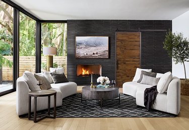Contemporary Barn Doors in room with Black brick wall with fireplace, rustic wood barn door, matching curved beige couches, black area rug, light wood floors.