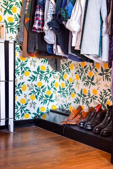 Closet floor with yellow and green floral wallpaper, hanging clothes, and shoe ledge.