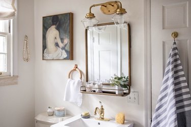 Bathroom with brass mirror, brass light fixture, striped towel, and painting.