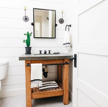 White powder room with wood vanity featuring concrete countertop and black hardware, black industrial mirror, and black and white striped towels. 