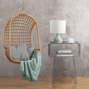 Image of a metal side table next to a chair swing. The chair swing is accessorized with a grey throw pillow and a teal blanket throw. The nightstand is accessorized with a teal and white lamp, a teacup, and books.