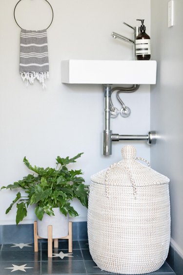 wall-mount white ceramic sink, blue floor tile with white star pattern, white planter with green plant, jute hamper, gray and white hand towel hung up