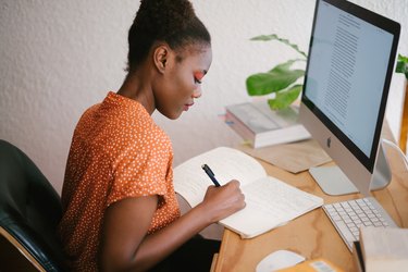 Black woman writing in notebook in front of apple desktop computer