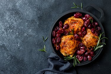 Roasted chicken legs with red grapes on pan over black slate stone background