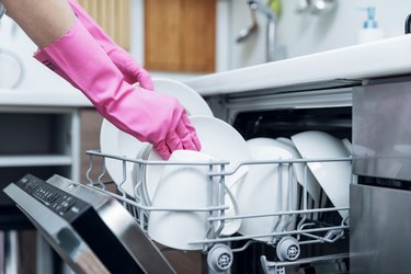 Woman wearing pink dishwashing gloves taking out clean dishes from dishwasher at home kitchen