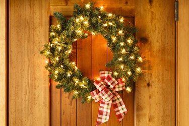 Beautiful lighted evergreen wreath with plaid fabric ribbon bow and lights hanging on wooden front door of home background. Simple, rustic country style Christmas holiday home decorations.