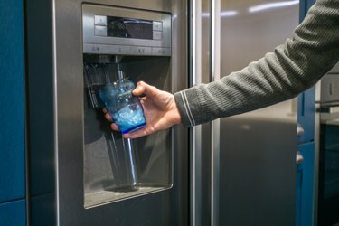 Male hand is pouring cold water and ice cubes from dispenser of home fridge.