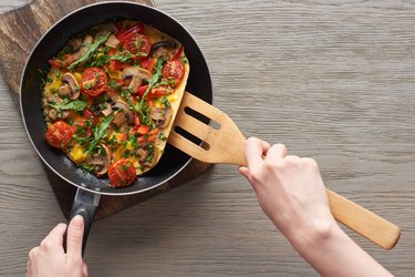cropped view of woman cooking omelet with mushrooms, tomatoes and greens on frying pan with wooden shovel