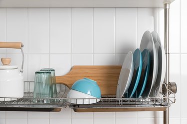 Clean dishes drying on metal dish rack on light background. Kitchen utensils and dishware on wooden shelf. Kitchen interior background.Text space.