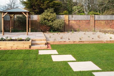 New stepping stones leading to steps and a raised patio with a wooden gazebo, next to a gravel stone planting area. The new turf has a brick mowing strip.