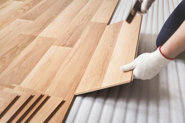 Installing laminated floor, man hand in white glove holding wooden tile and using hammer, over white foam base layer