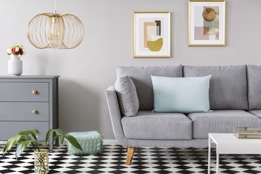 Light blue pillow on grey couch in bright living room interior with checkerboard linoleum floor, fresh flowers in vase on cupboard two posters hanging on the wall and gold lamp.
