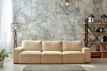 Modern interior of living room with gray walls, beige sofa, floor lamp and chandelier, light carpet shelf with frames and jars, flower in a pot