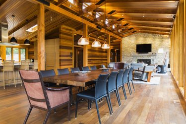 Wooden dining table with rawhide armchairs, black leather chairs and illuminated industrial style copper with frosted glass pendant lighting fixtures in dining area of great room with Ipe wood floor plus view of living room area with natural stone firepl