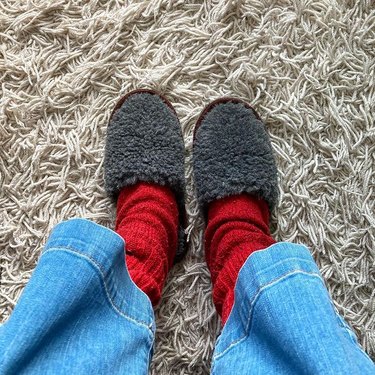 person wearing grey ikea fegen slippers with red socks and jeans on white stringy carpet