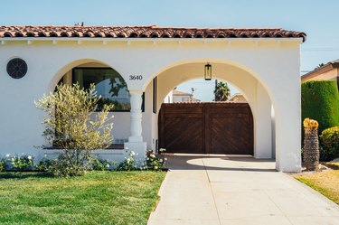 A white Spanish-style home with a red-tiled roof; curved archways cover a wooden gate and a lush green yard is in front