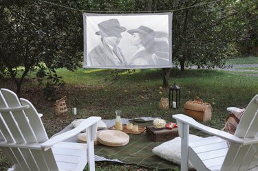 DIY portable projector screen hanging outside between two trees with chairs, blankets and snacks set in front