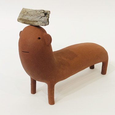 Handcrafted rust-colored ceramic sculpture of imaginary animal with piece of wood on its head by Godeleine de Rosamel.