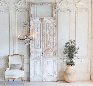 White weathered double doors with frame in white room next to white upholstered chair and potted planr