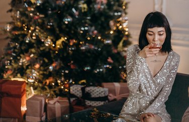 woman on sofa in sparkly dress sipping on cocktail with christmas trees and presents in background