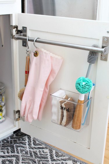under sink storage with towel bar with pink gloves and scrub brushes hanging on white cabinet door