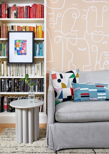 How To Organize a Bookshelf in Living room with pale pink patterned wallpaper, color coded book shelves, gray couch, white modern end table, colorful throw pillows, art.