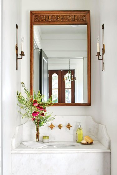 French country bathroom sink with marble vanity, candle sconces, vintage mirror, wall mounted faucet, flowers, soaps.