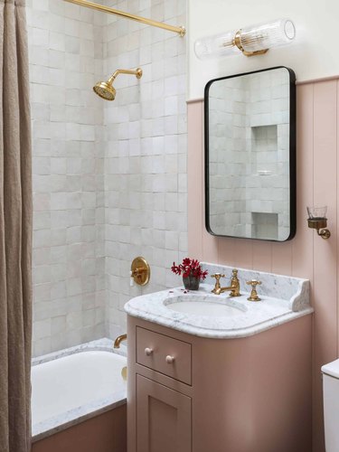 French country bathroom sink in bathroom with rose pink vanity, white tiles, marble counter, under mount sink, brass faucet and shower head.