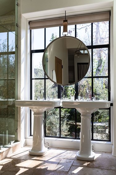 French country bathroom sinks with round mirror, large window, stone floors.
