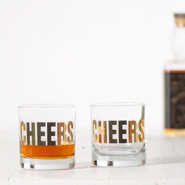 two glasses with "cheers" in gold and a bottle out of focus in the background