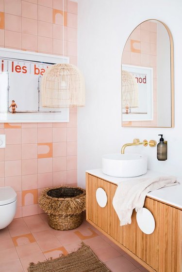 Pink bathroom with brass Wall-Mounted Bathroom Faucet, wood vanity, bowl sink, basket, cane pendant lamp, mirro.