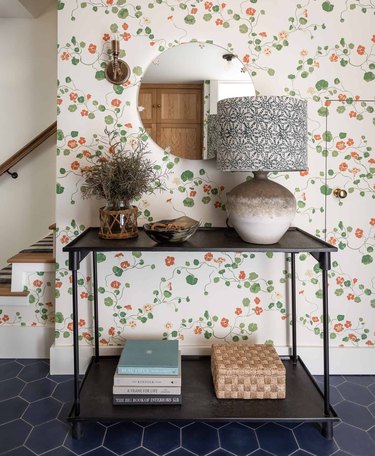 Entryway with floral wallpaper, console table, lamp, books, mirror.