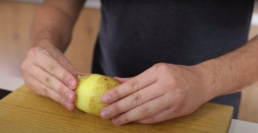 person peeling skin off of potato on wood cutting board