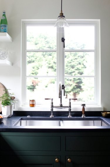 Best kitchen sink materials Kitchen sink with stainless faucet, window, pendant light, open shelves, plant, black counters, green cabinets.