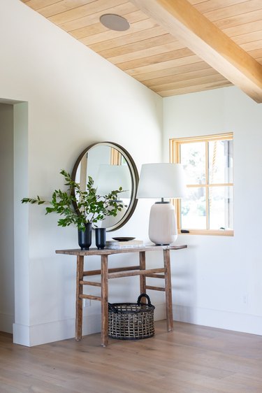 Entryway with table, round mirror, big white table lamp, plants, basket, window, wood floors.
