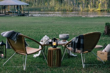 Two lawn chairs in the foreground with a table set up with a s'mores station; a lush green lawn with a river in the background