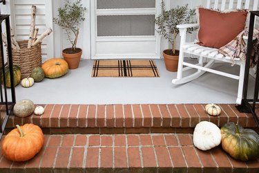 Plaid doormat, firewood basket, potted olive trees and pumpkins on fall porch