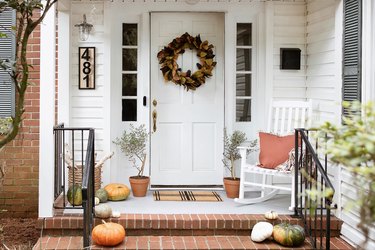 Fall porch with wreath, pumpkins, rocking chair, house numbers, and firewood stored in basket