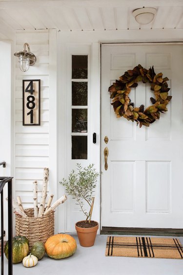 Faux magnolia wreath hung on door with plaid doormat, DIY house numbers plaque, industrial porch light, firewood basket, and pumpkins on fall porch