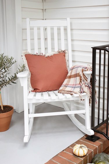 White rocking chair with tan tartan plaid blanket and rust-colored fringe pillow on porch