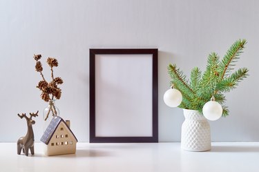 Mock up black poster frame with christmas decoration in home interior, scandinavian style. Christmas little house and deer, green fir branches in a vase on a white table